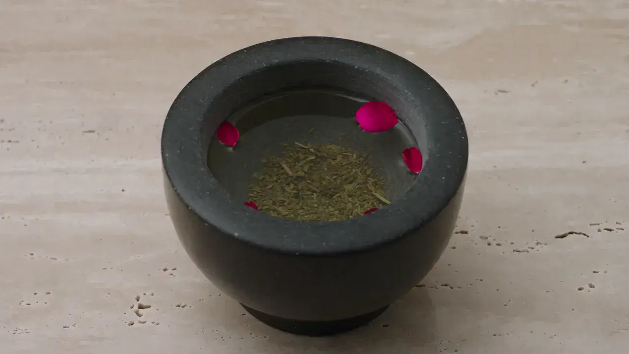 A dark stone mortar holding dried herbs and floating rose petals on pale travertine